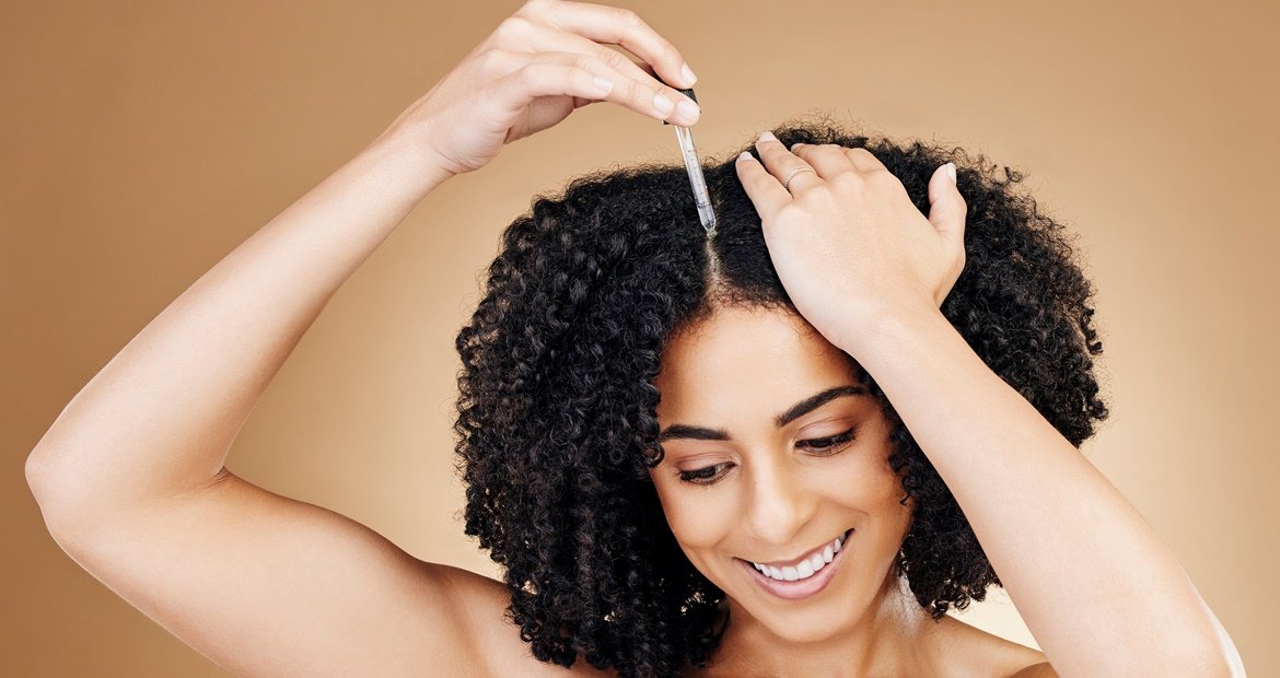 Mulher sorrindo enquanto passa óleo de girassol em seu cabelo cacheado.